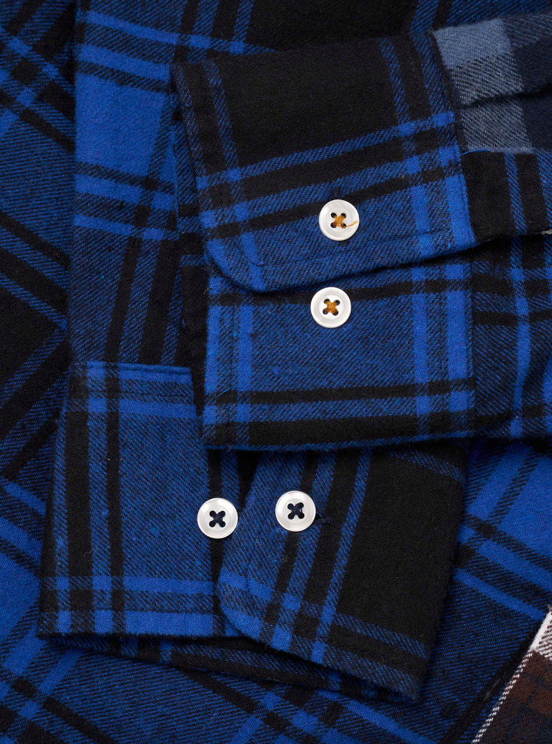 Close-up flat lay of a blue and black patchwork check flannel shirt. Two cuffs are visible, each fastened with white four-hole buttons. The upper cuff has one button, the lower cuff has two. Brushed twill fabric shows bold cobalt and black tartan checks across contrasting patchwork panels.