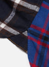 Close-up flat lay of a blue patchwork checks shirt on a white surface. Two contrasting tartan panels are visible: a dark navy, brown and white check on the left, and a bright blue, red and black tartan on the right. A seam joins the panels with visible mustard yellow stitching. Soft, brushed flannel texture throughout.
