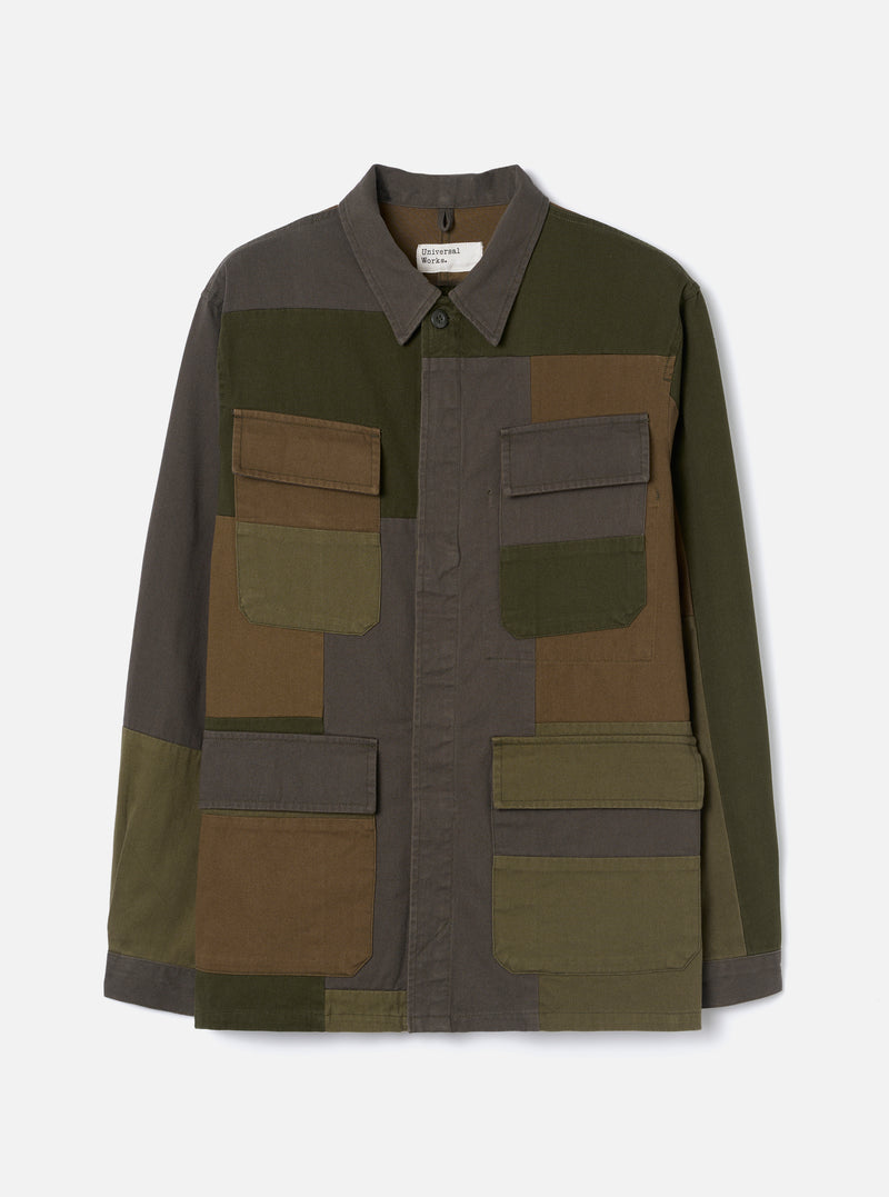 Olive patchwork twill fatigue jacket laid flat on a light grey studio backdrop. Patchwork panels in olive, khaki, charcoal and brown cover the body. Point collar, single button placket and a Universal Works woven label at the chest. Four large patch pockets with buttoned flaps, two chest and two lower. Long sleeves with plain cuffs.