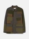 Olive patchwork twill fatigue jacket laid flat on a light grey studio backdrop. Patchwork panels in olive, khaki, charcoal and brown cover the body. Point collar, single button placket and a Universal Works woven label at the chest. Four large patch pockets with buttoned flaps, two chest and two lower. Long sleeves with plain cuffs.