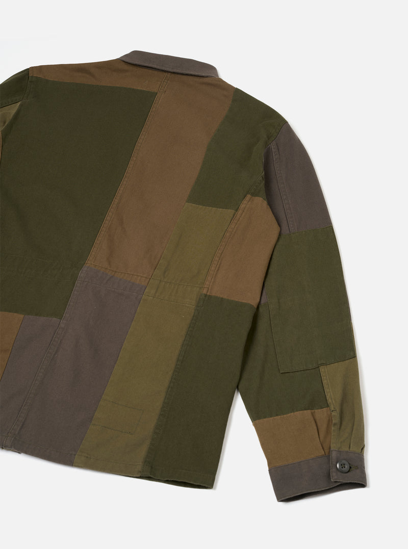 Back view of an olive patchwork twill fatigue jacket on a plain light grey studio backdrop. The jacket features large colour-blocked panels in olive, khaki brown and charcoal, sewn together with visible seams. A small patch pocket sits on the lower sleeve. The cuff is charcoal with a single dark button.