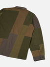 Back view of an olive patchwork twill fatigue jacket on a plain light grey studio backdrop. The jacket features large colour-blocked panels in olive, khaki brown and charcoal, sewn together with visible seams. A small patch pocket sits on the lower sleeve. The cuff is charcoal with a single dark button.