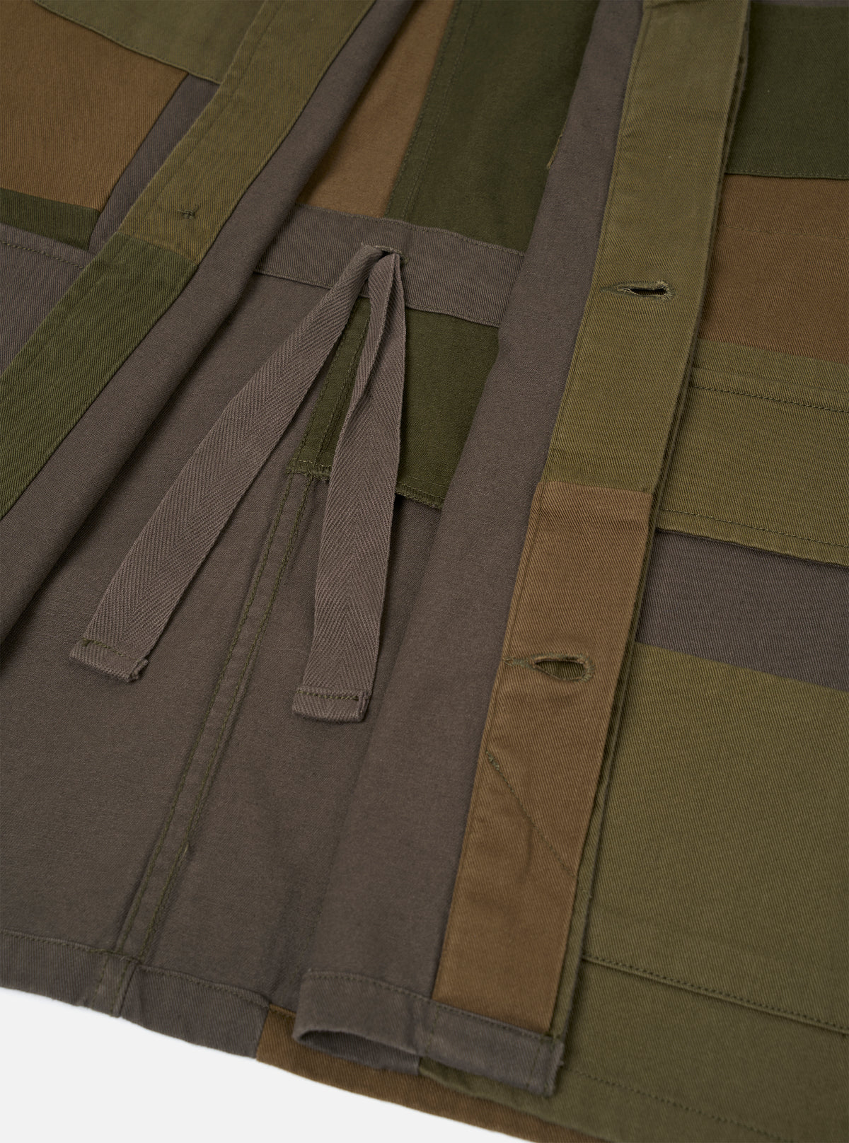 Close-up flat lay of an olive patchwork twill fatigue jacket showing the front placket and lower body. Patchwork panels in olive, khaki brown and charcoal grey are visible. A herringbone twill tape tie is knotted at the waist opening. Two vertical buttonholes are visible on the right placket. Stitched hem detail shows at the bottom edge.