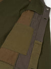 Close-up detail of an olive patchwork twill fatigue jacket on a white studio backdrop. The shot highlights the front placket and chest area, showing patchwork panels in olive, brown, and dark grey twill. A large patch pocket sits at centre with topstitched edges. The placket features distressed buttonholes with raw frayed edges between panels.
