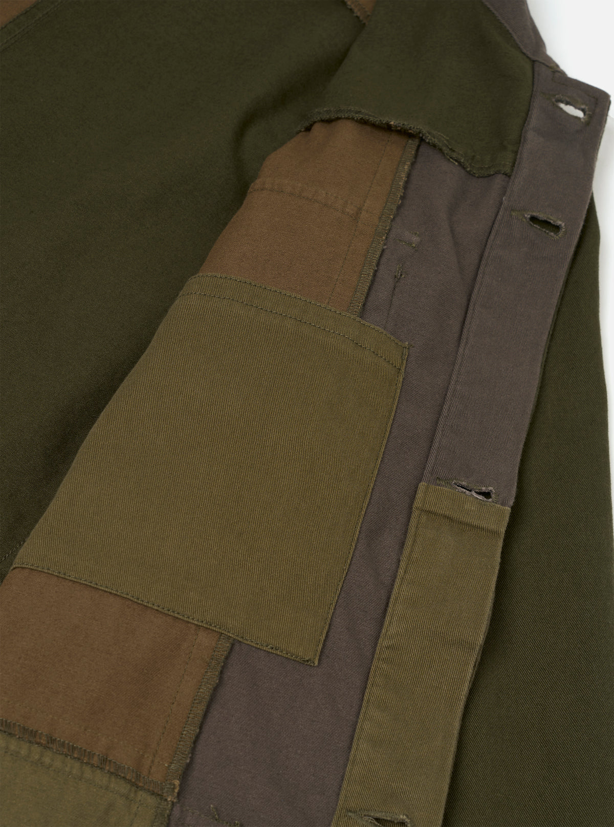 Close-up detail of an olive patchwork twill fatigue jacket on a white studio backdrop. The shot highlights the front placket and chest area, showing patchwork panels in olive, brown, and dark grey twill. A large patch pocket sits at centre with topstitched edges. The placket features distressed buttonholes with raw frayed edges between panels.