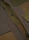 Close-up detail of an olive patchwork twill fatigue jacket laid flat. The button placket shows two dark four-hole buttons on a twill facing. Patchwork panels in olive, khaki, and brown tones are visible across the body. Patch pockets with raw-edge detailing sit on the chest and lower front. Plain dark studio backdrop.