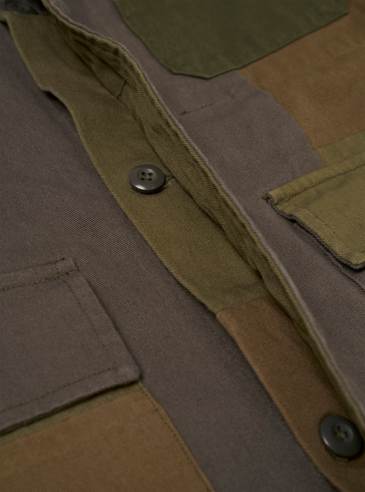 Close-up detail of an olive patchwork twill fatigue jacket laid flat. The button placket shows two dark four-hole buttons on a twill facing. Patchwork panels in olive, khaki, and brown tones are visible across the body. Patch pockets with raw-edge detailing sit on the chest and lower front. Plain dark studio backdrop.