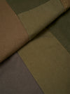 Close-up detail of the olive patchwork twill fatigue jacket laid flat, showing colour-blocked panels in olive, tan, dark khaki and charcoal. A folded seam runs diagonally across the centre, highlighting the contrast stitching where panels meet. The twill weave texture is clearly visible across all sections.