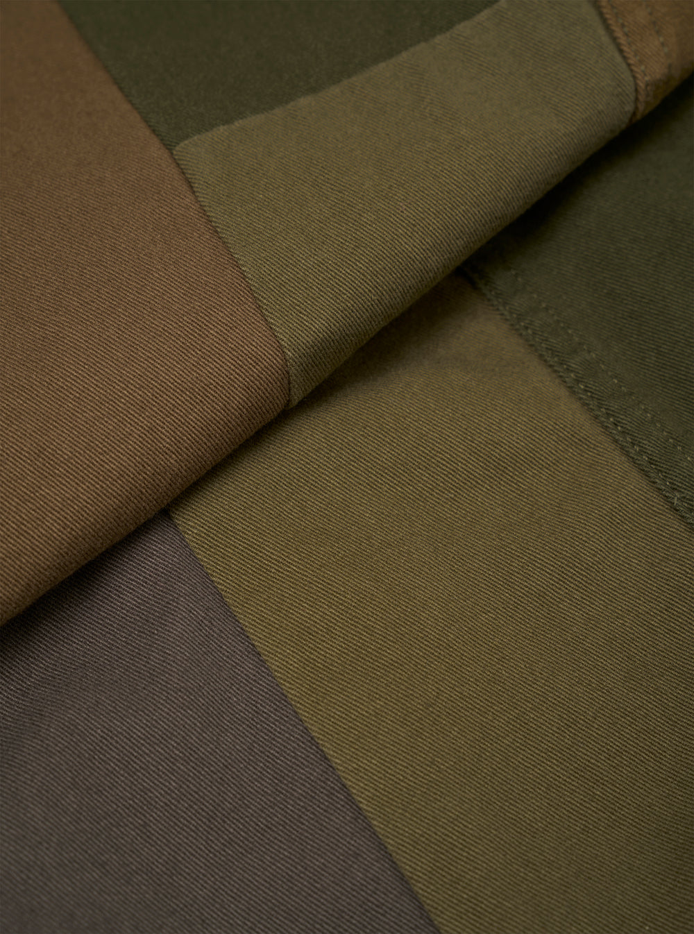 Close-up detail of the olive patchwork twill fatigue jacket laid flat, showing colour-blocked panels in olive, tan, dark khaki and charcoal. A folded seam runs diagonally across the centre, highlighting the contrast stitching where panels meet. The twill weave texture is clearly visible across all sections.