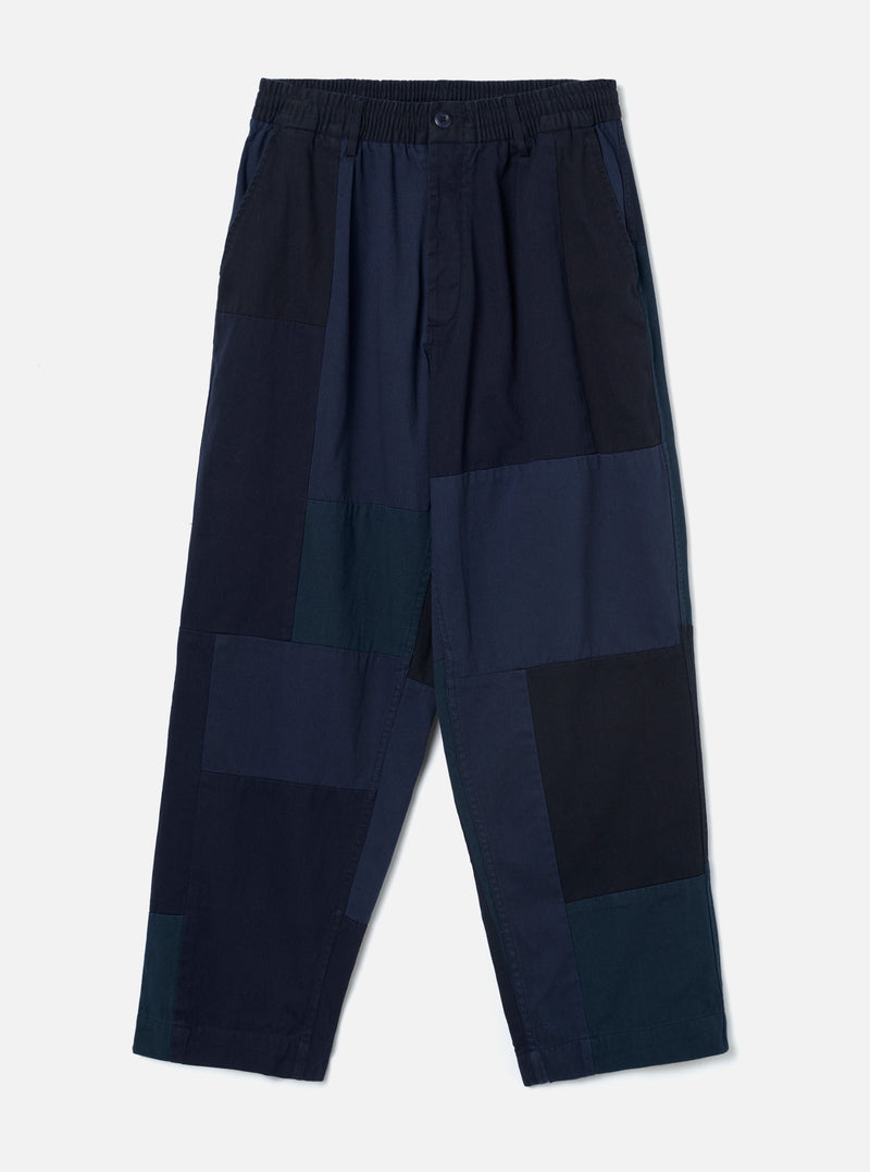Navy patchwork twill Oxford trousers laid flat on a plain light grey backdrop. Wide-leg cut with an elasticated waistband, single centre button, and slant side pockets. The body features large irregular patchwork panels in varying shades of navy, teal and black woven twill. Full-length leg with an unfinished hem.