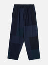 Navy patchwork twill Oxford trousers laid flat on a plain light grey backdrop. Wide-leg cut with an elasticated waistband, single centre button, and slant side pockets. The body features large irregular patchwork panels in varying shades of navy, teal and black woven twill. Full-length leg with an unfinished hem.