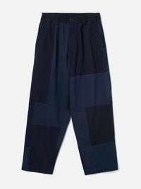 Navy patchwork twill Oxford trousers laid flat on a plain light grey backdrop. Wide-leg cut with an elasticated waistband, single centre button, and slant side pockets. The body features large irregular patchwork panels in varying shades of navy, teal and black woven twill. Full-length leg with an unfinished hem.