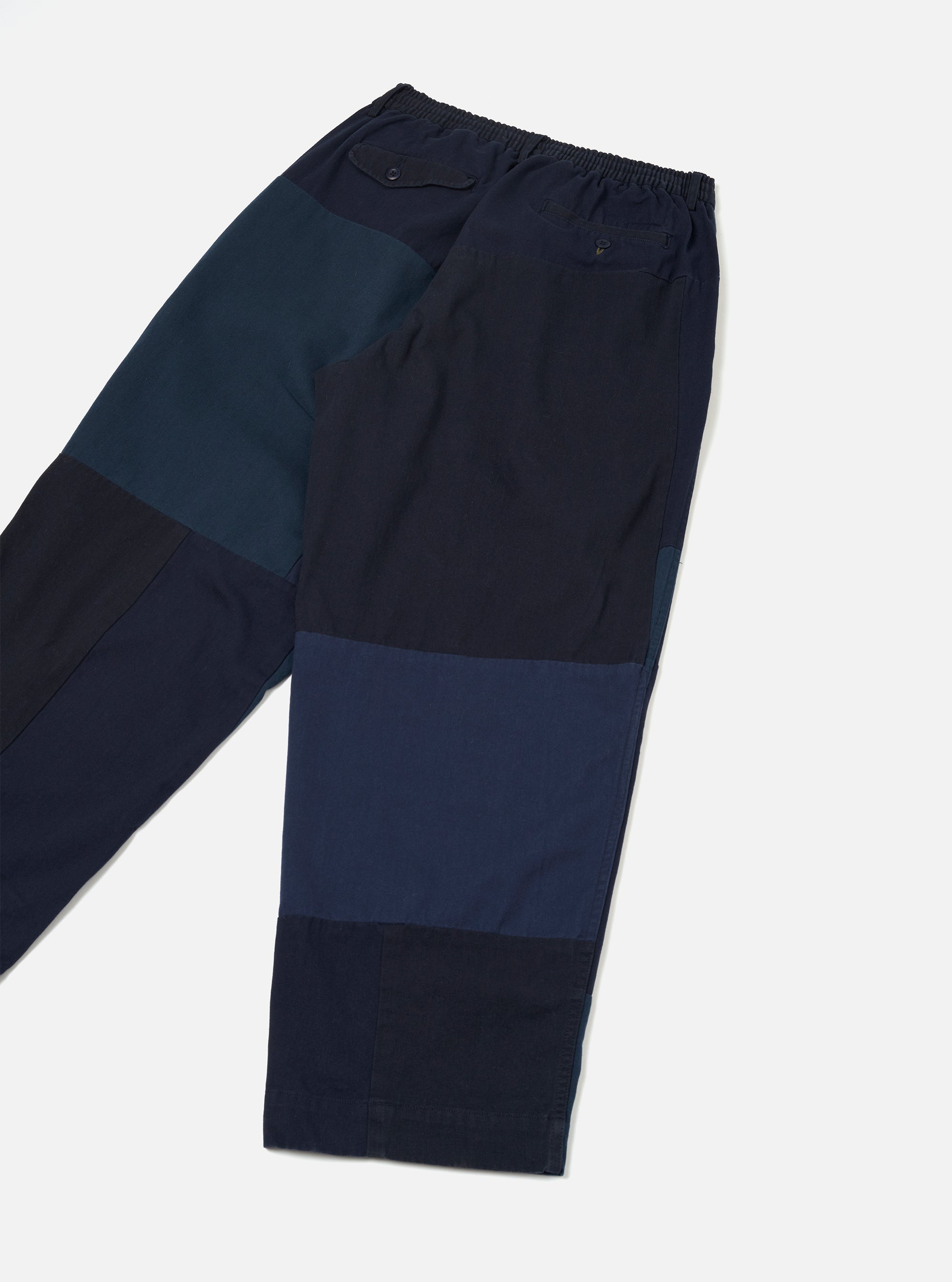 Navy patchwork twill Oxford trousers laid flat on a light grey surface, cropped above the knee. Elasticated waistband with a small flap pocket and a rear welt pocket with button. Patchwork panels in navy, dark teal, and mid-blue tones. Woven twill texture visible throughout.