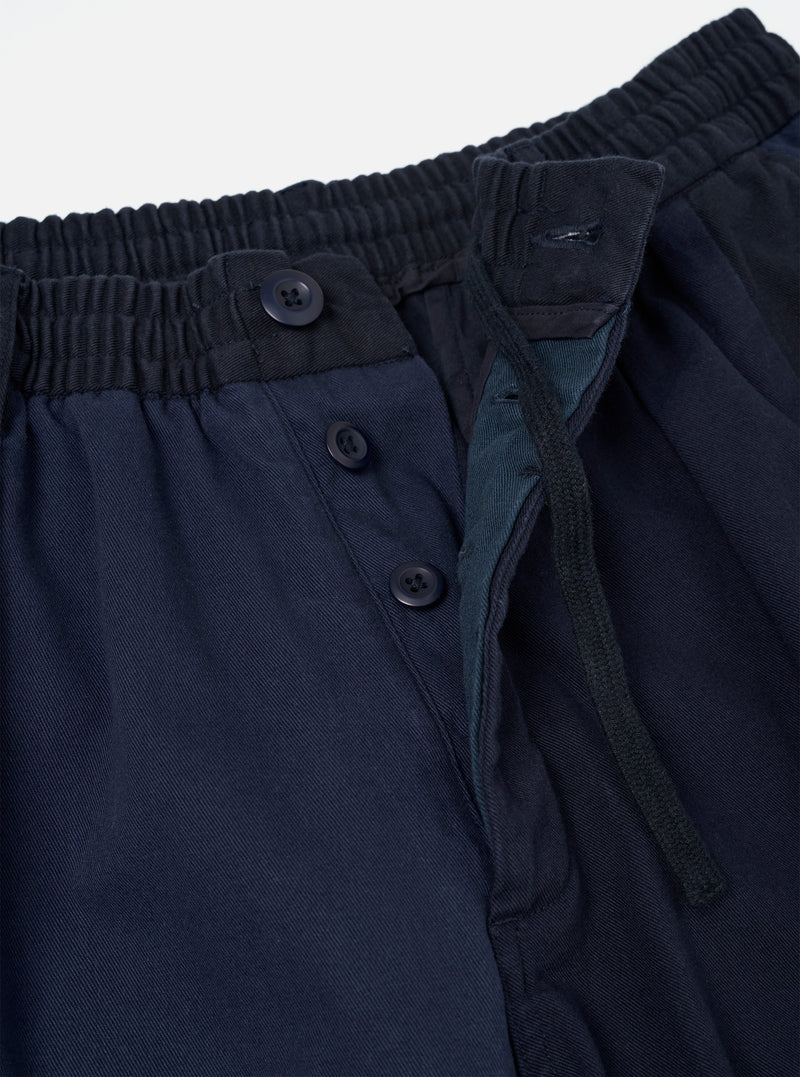 Close-up of the waistband and fly area of navy twill Oxford trousers. The elasticated waistband shows fine ribbed gathering. A button fly features three dark navy buttons on a placket, with a teal-toned inner fly lining visible. The twill weave is clear across the fabric. Plain white studio backdrop.