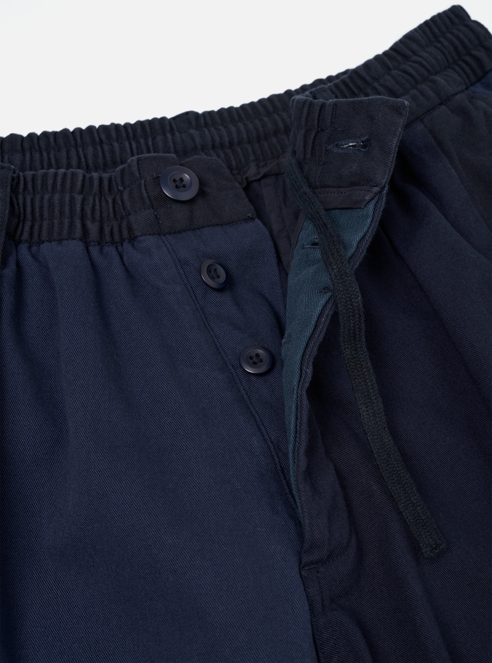 Close-up of the waistband and fly area of navy twill Oxford trousers. The elasticated waistband shows fine ribbed gathering. A button fly features three dark navy buttons on a placket, with a teal-toned inner fly lining visible. The twill weave is clear across the fabric. Plain white studio backdrop.