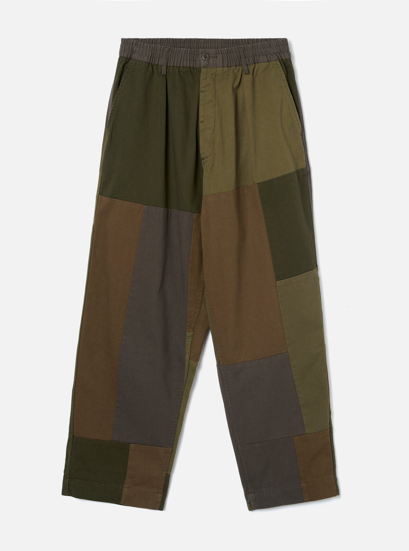 Olive patchwork twill Oxford pant laid flat on a plain light grey background. The wide-leg trouser features an elasticated waistband with a single button fastening, side pockets, and a full-length hem. Bold colour-block panels in olive, dark khaki, brown and charcoal grey are stitched together across both legs in a patchwork construction.