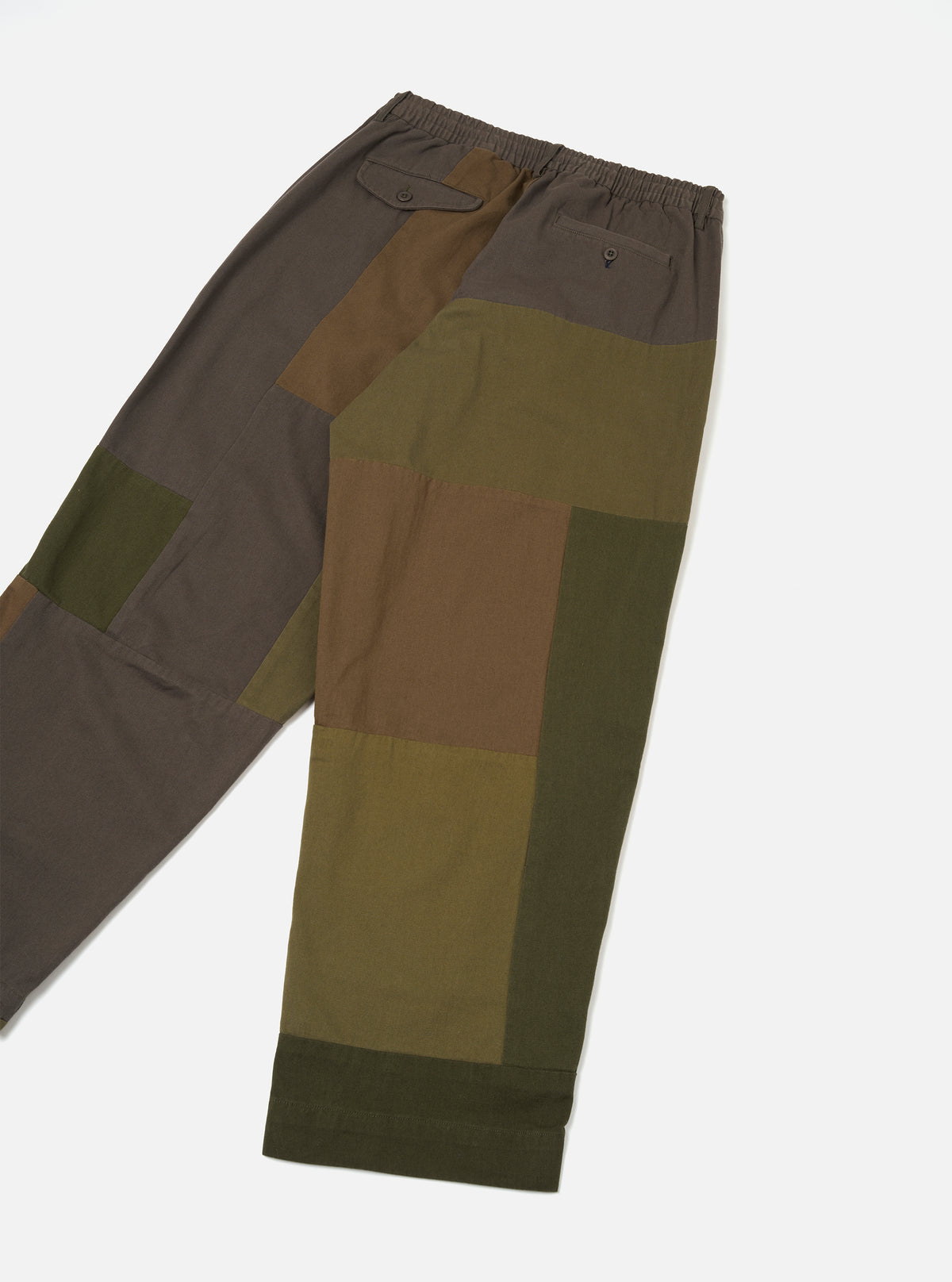 Olive patchwork twill Oxford trousers laid flat on a white studio surface. The fabric features large colour-blocked panels in olive, khaki, brown and dark grey. An elasticated waistband sits at the top, with a buttoned flap pocket on the left and a small button-through pocket on the right. Cropped legs with cuffed hems.