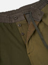 Close-up detail of olive patchwork twill trousers on a plain light grey studio backdrop. Shot shows a ribbed dark brown elasticated waistband with a braided olive drawcord. The open fly reveals three dark brown four-hole buttons on a placket, with contrast olive twill lining visible beneath.