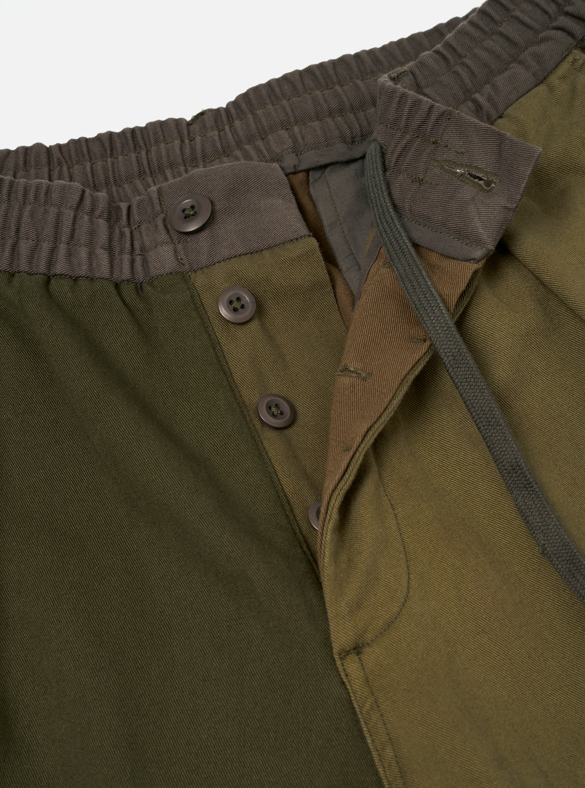 Close-up detail of olive patchwork twill trousers on a plain light grey studio backdrop. Shot shows a ribbed dark brown elasticated waistband with a braided olive drawcord. The open fly reveals three dark brown four-hole buttons on a placket, with contrast olive twill lining visible beneath.