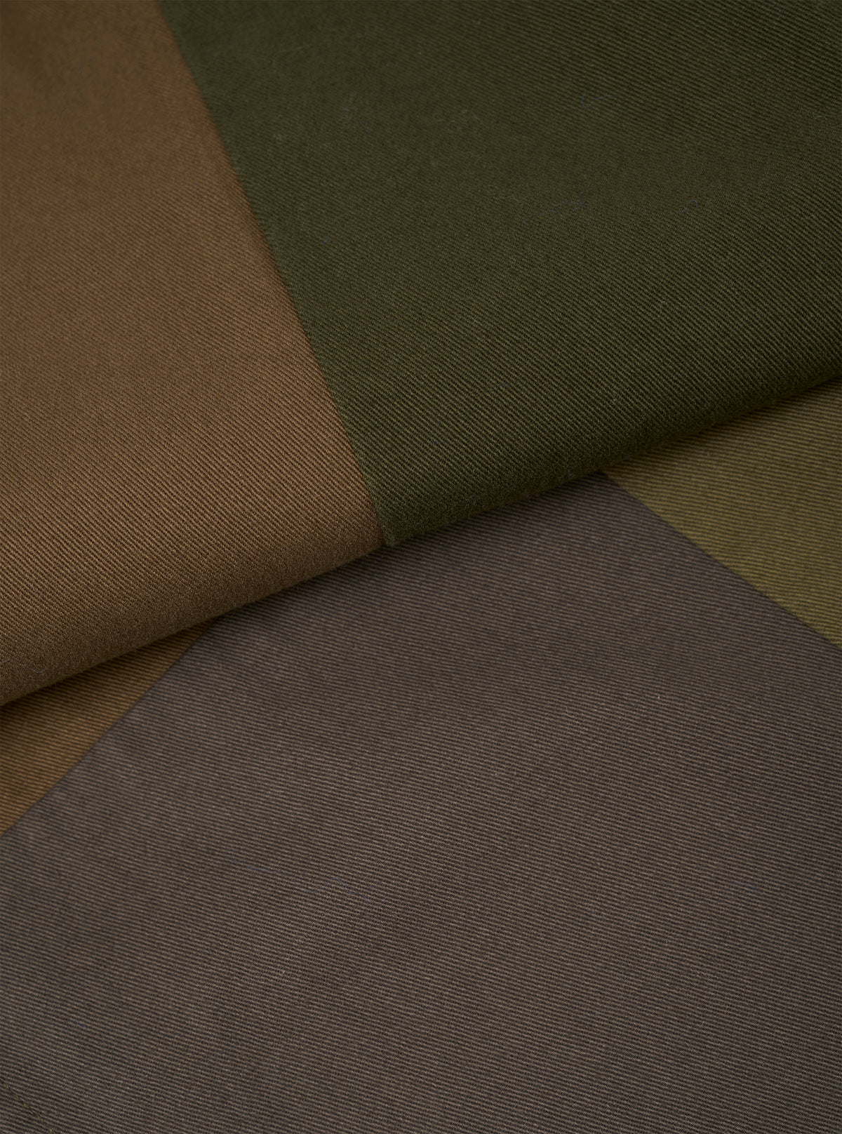 Close-up flat lay showing four colour swatches of twill fabric arranged in diagonal panels: tan, dark olive, muted khaki, and grey-brown. The fine diagonal weave of the twill is clearly visible across all panels, illustrating the fabric options available for the Olive Patchwork Twill Oxford Pant.