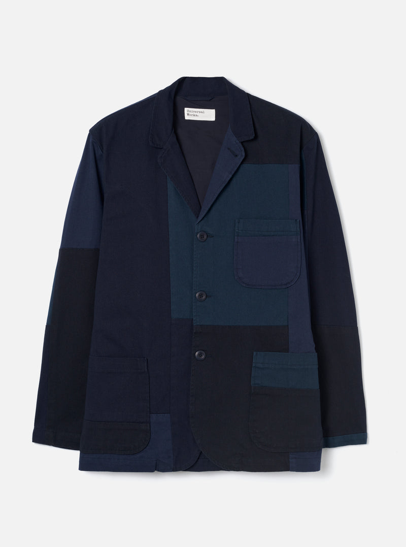 Navy patchwork blazer-style jacket laid flat on a light grey studio backdrop. Notched lapels, three dark buttons, a chest patch pocket and two large lower patch pockets. Patchwork panels in navy, teal and black twill fabrics. Universal Works woven label at the collar. Full length sleeves, straight hem.