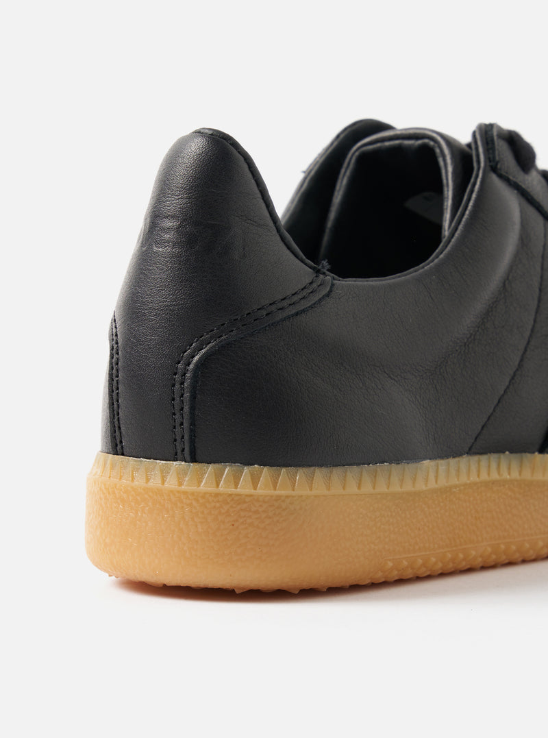 Close-up of the Novesta for Universal Works Black Leather German Army Trainer. Features black leather upper and gum sole. Shot on a white background.