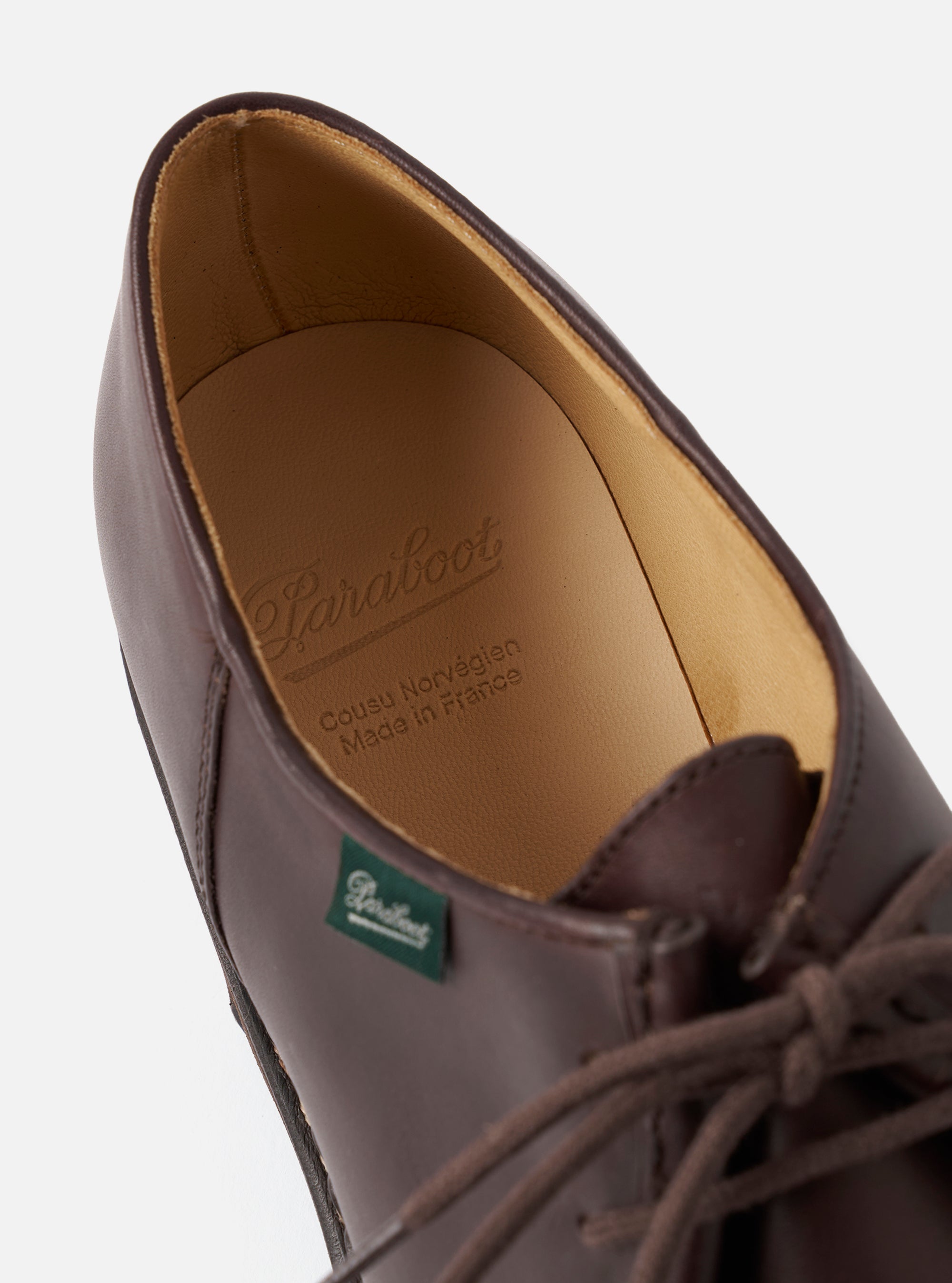 Close-up of a Paraboot cafe brown leather derby shoe on a light grey studio backdrop. The tongue is folded back to reveal the tan leather insole, embossed with the Paraboot logo and "Cousu Norvégien, Made in France." A green Paraboot branded label is visible on the tongue. Round brown laces are partially visible.