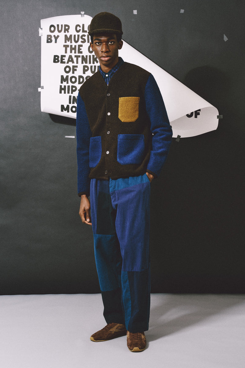 A model stands in a studio wearing a dark brown and blue patchwork corduroy jacket with matching patchwork trousers, a dark cap, and brown shoes. A large white paper cutout with text is held behind his shoulder against a dark gray backdrop.