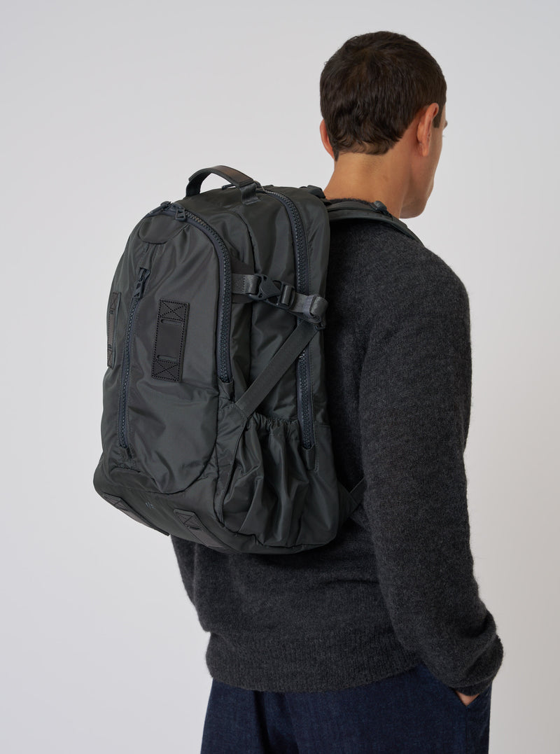 A model wears the F/CE.® Grey Recycled Nylon 420 re/cor Travel Backpack. The grey nylon backpack is on a model in a grey sweater, against a white backdrop.