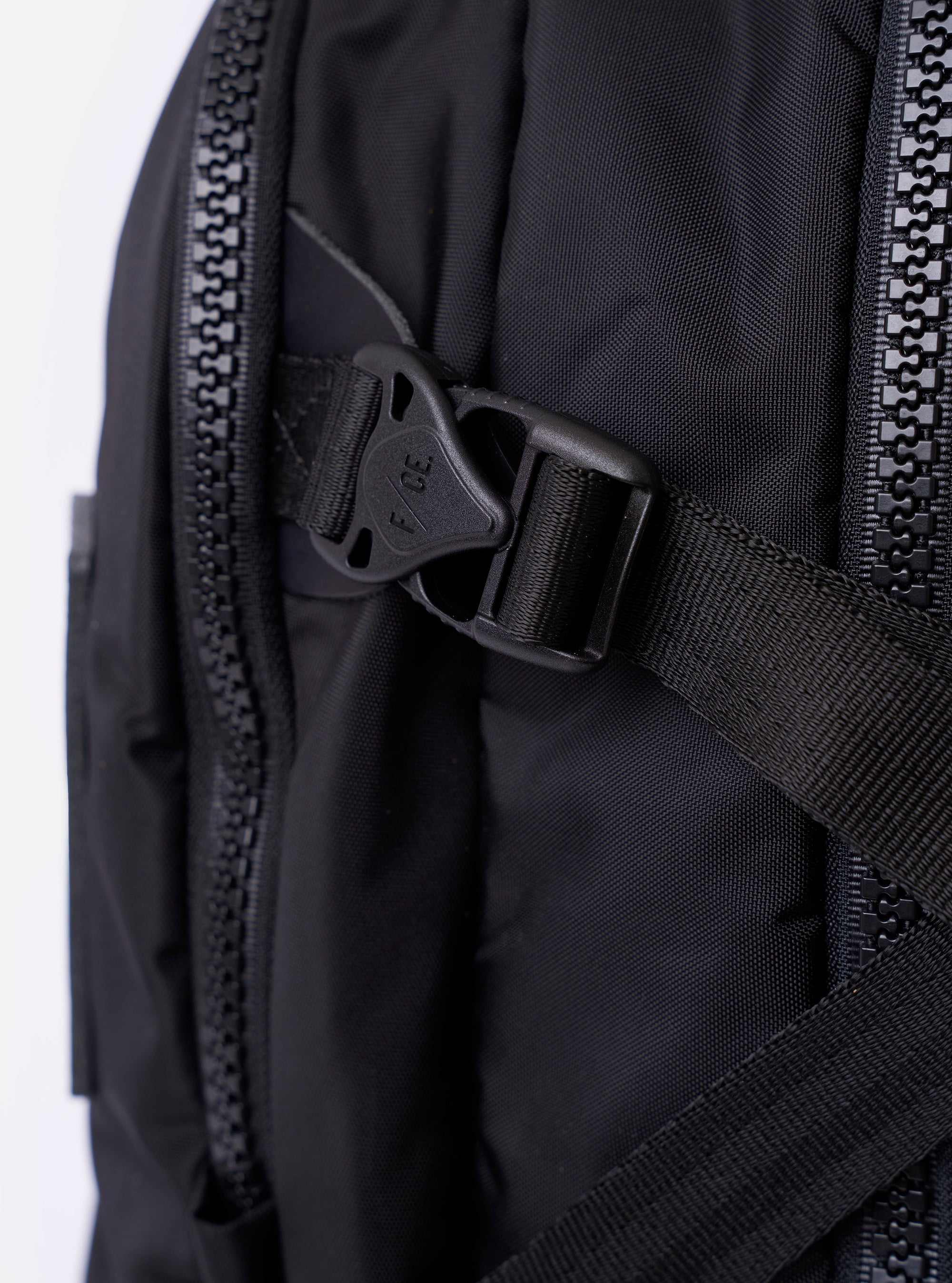 The F/CE.® Black Recycled Nylon 420 re/cor Travel Backpack is shown from a rear angle against a plain white background. The image highlights the padded, ergonomic shoulder straps with the Mounting T System, a mesh-padded back panel for comfort, and a top carry handle. The backpack is made from a sleek, black recycled nylon fabric.