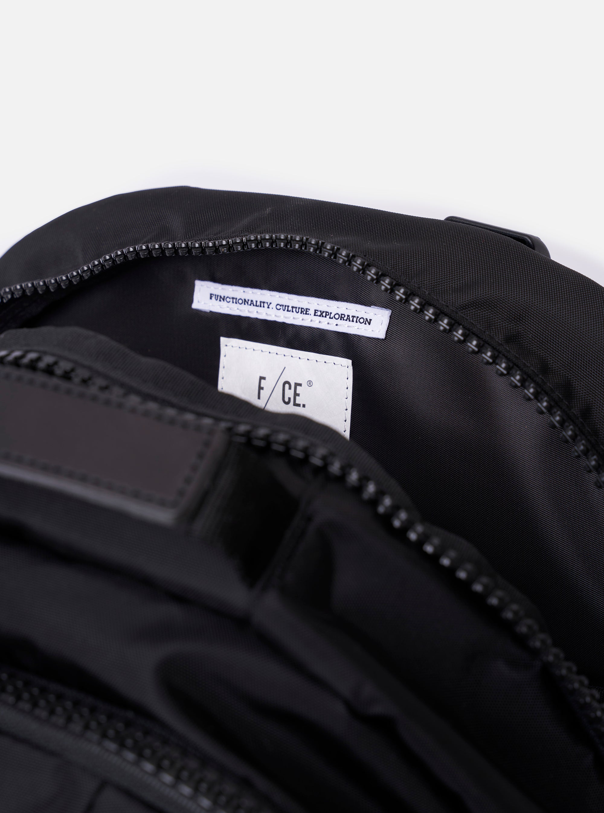 The F/CE.® Black Recycled Nylon 420 re/cor Travel Backpack is shown from the back as a flat lay against a plain white background. The image highlights the ergonomic, curved shoulder straps featuring the Mounting T System and a small embroidered logo. The back panel is padded with a smooth, black material.