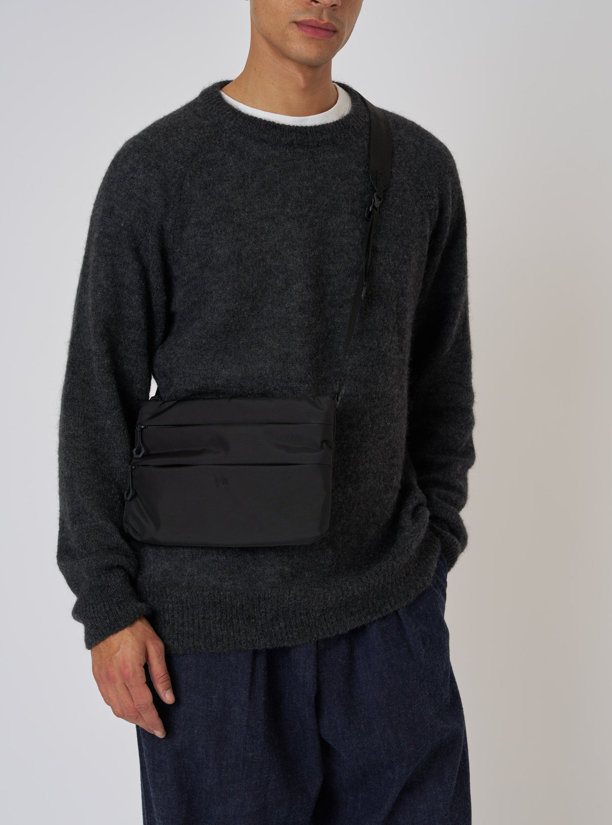 A model showcases the F/CE.® Black 420 re/cor Sacoche against a plain backdrop. The black bag is worn across the body with a dark sweater and blue pants.