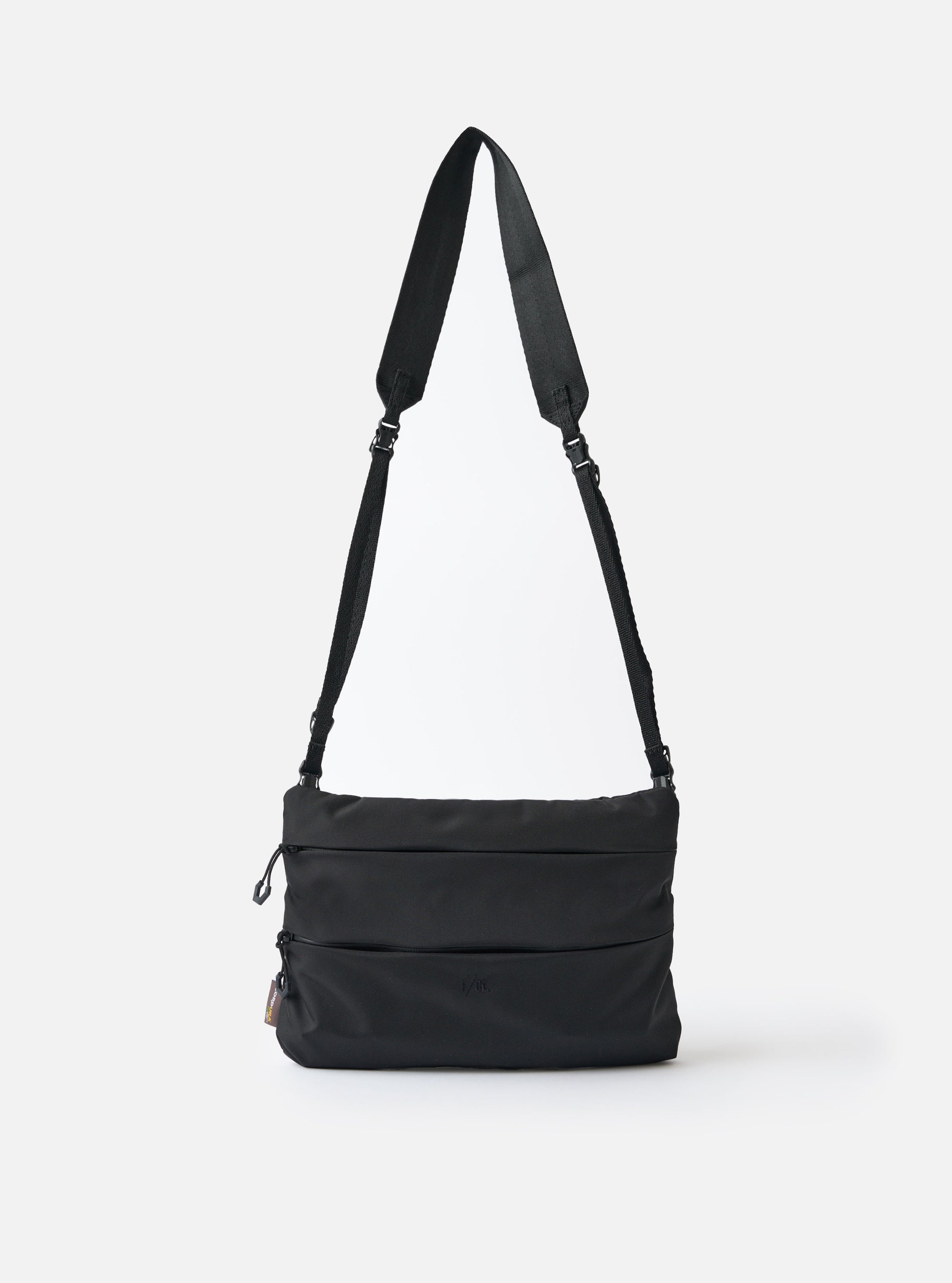 The F/CE.® Black 420 re/cor Sacoche bag is displayed on a white background. The black bag has a long black strap. The bag has a rectangular shape with horizontal stitched lines.