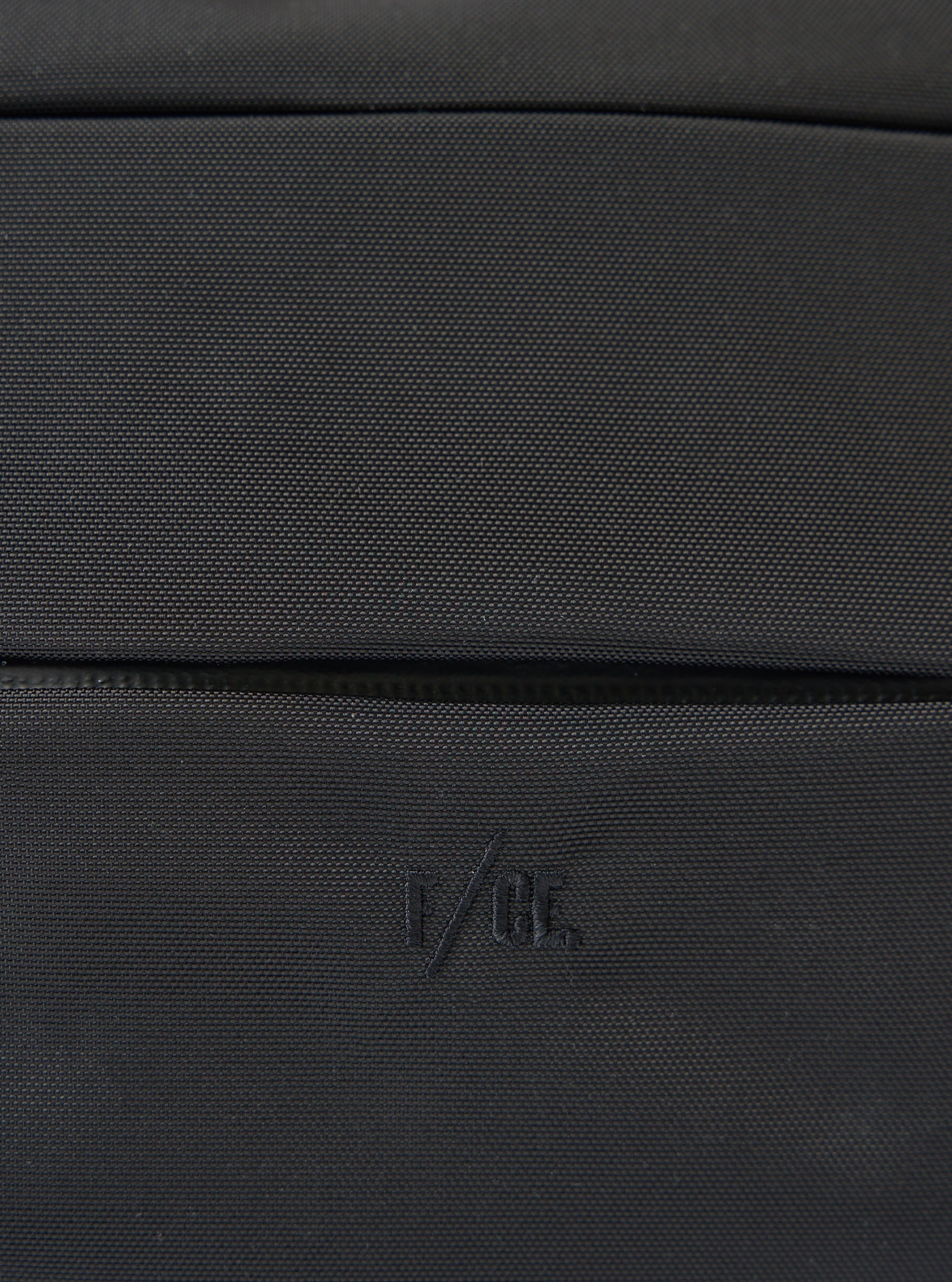 Close-up of the F/CE.® Black 420 re/cor Sacoche. The black textured material is visible. The F/CE logo is embroidered on the bottom. Shot on a flat surface.