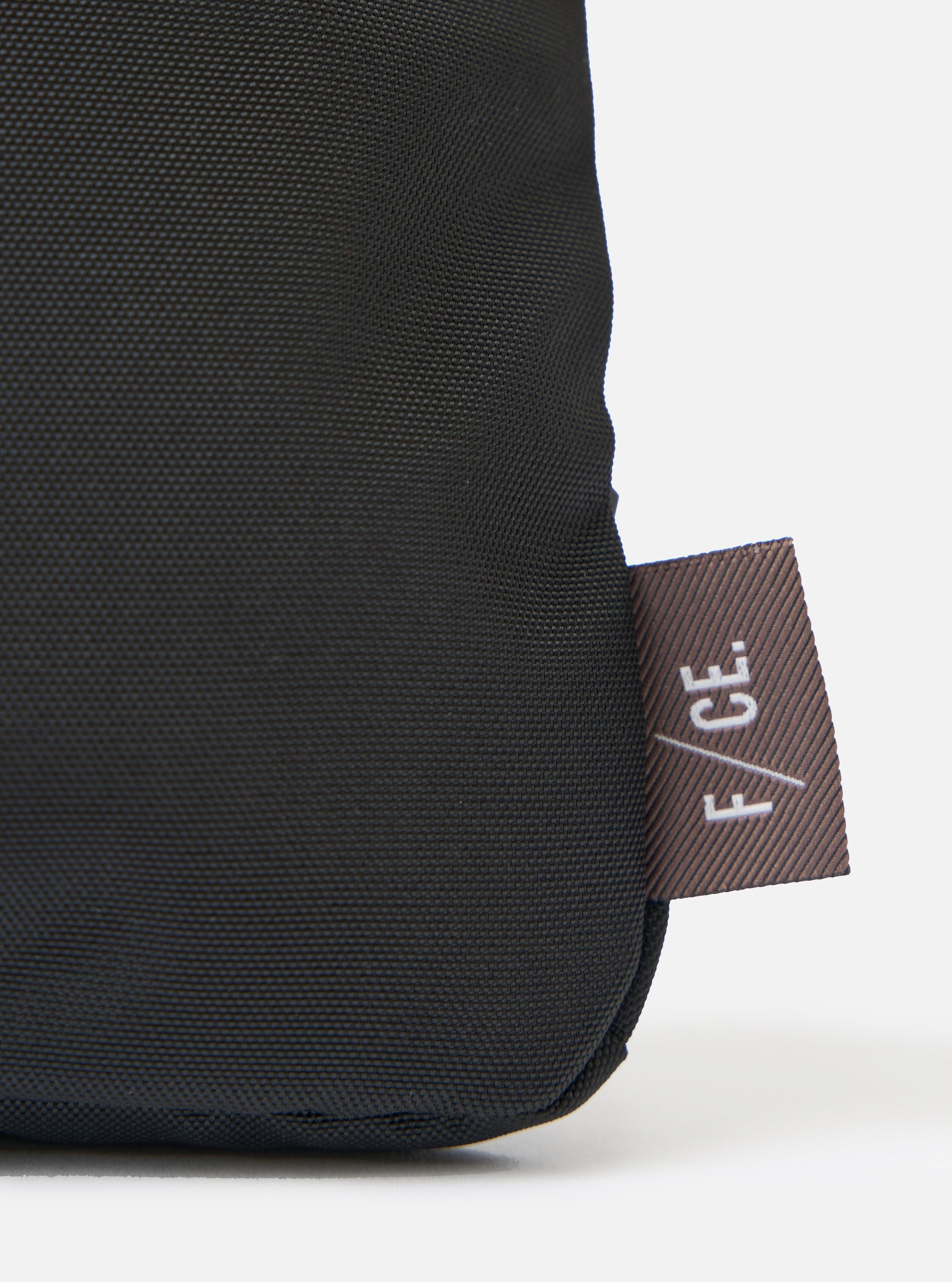 Close up of the F/CE.® Black 420 re/cor Sacoche showing the black textured material and the brown F/CE. tag on a white background.
