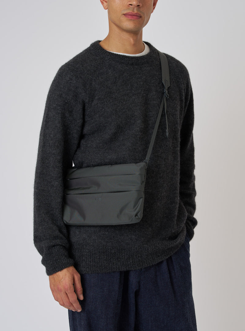 A model showcases the F/CE.® Grey 420 re/cor Sacoche. The grey bag is worn across the body. The model wears a dark grey sweater and blue pants. Simple studio background.