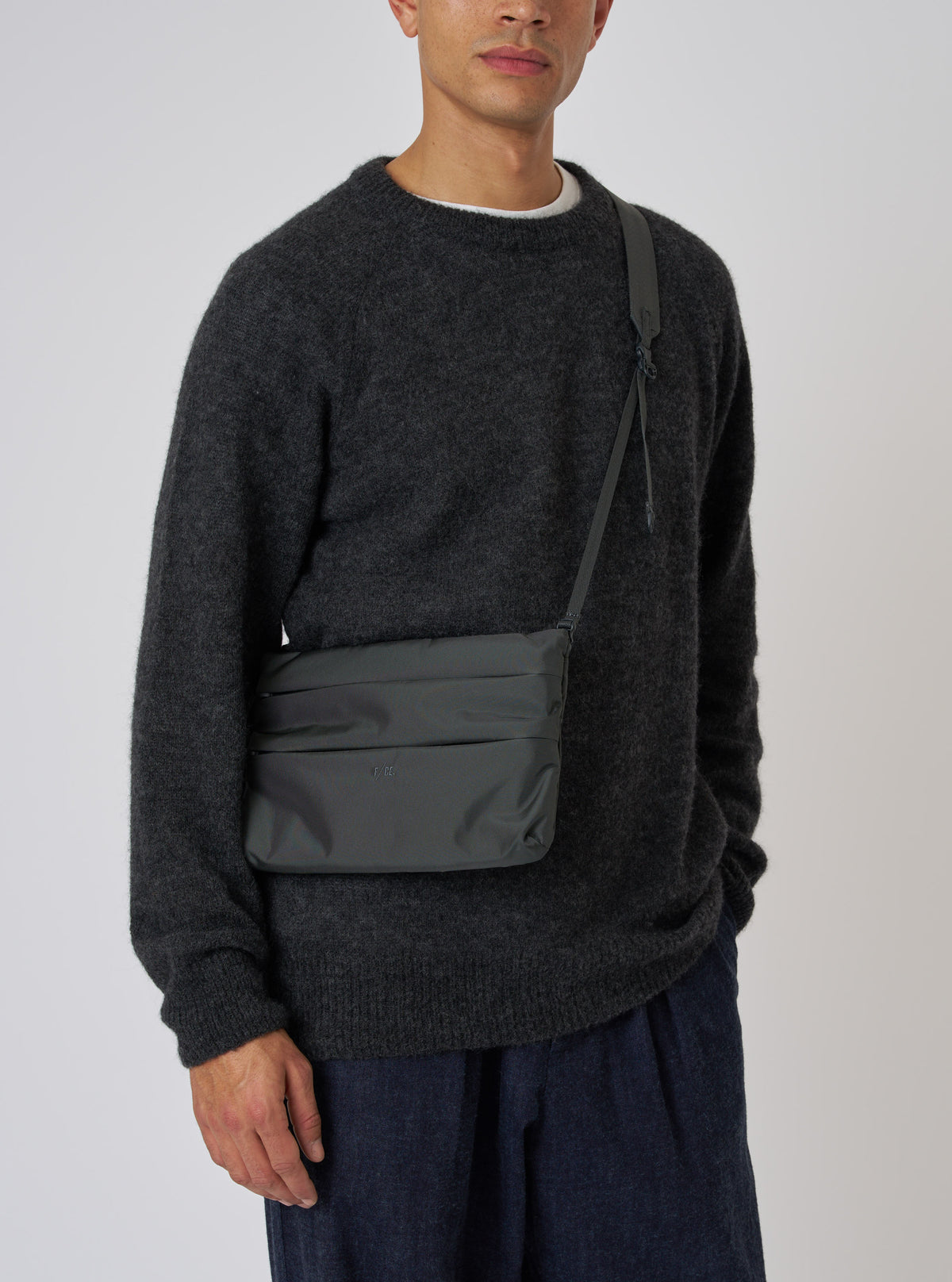 A model showcases the F/CE.® Grey 420 re/cor Sacoche. The grey bag is worn across the body. The model wears a dark grey sweater and blue pants. Simple studio background.