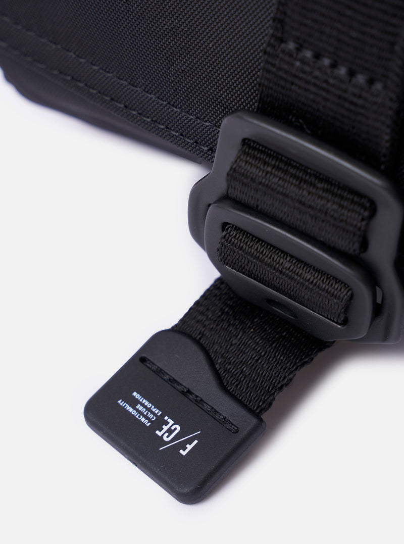 The F/CE.® Black Recycled Nylon 420 re/cor Holiday Sacoche is shown as a flat lay against a plain white background. The compact, rectangular travel bag features two zippered compartments, an adjustable and detachable shoulder strap with plastic buckles, and a small brand label on the front. The black recycled nylon fabric has a smooth, technical appearance.