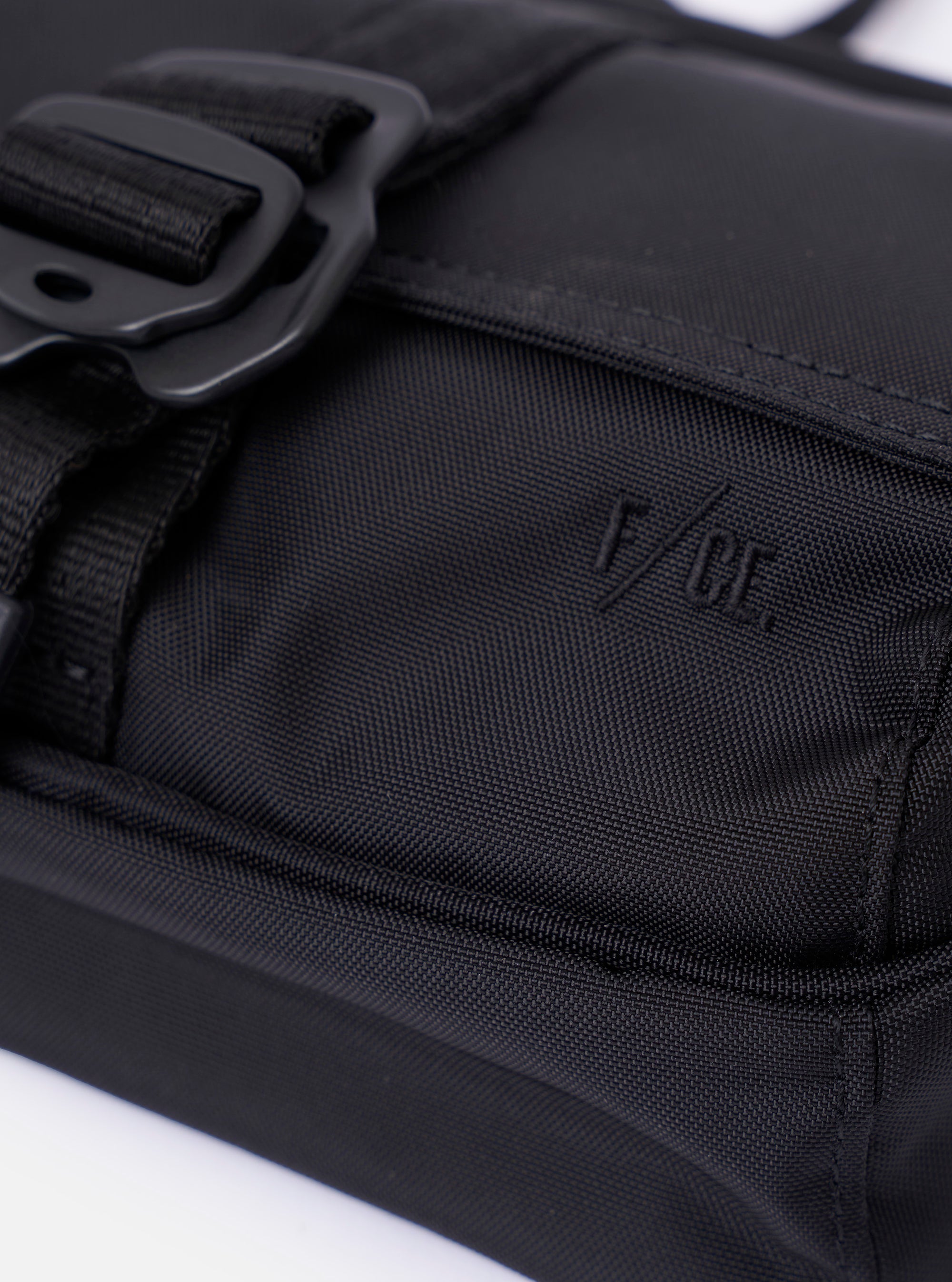 The F/CE.® Black Recycled Nylon 420 re/cor Holiday Sacoche is shown in a detail view as a flat lay against a plain white background. The image highlights the smooth black recycled nylon fabric, focusing on the two-compartment design and the clean stitching of the front pocket. A slim, adjustable black shoulder strap and a small embroidered logo are visible.