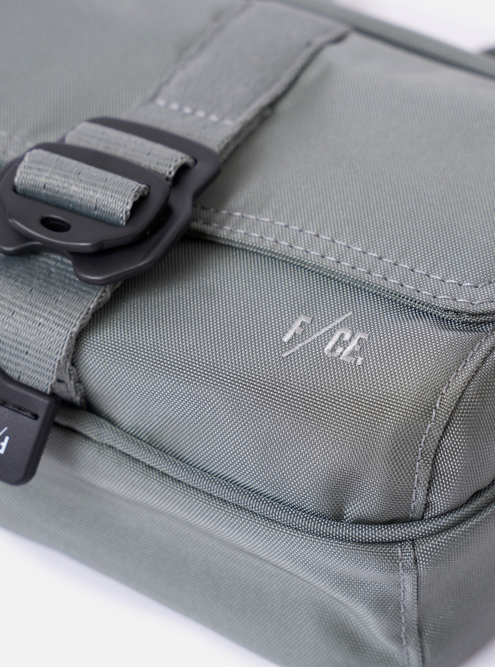 The F/CE.® Foliage Green Recycled Nylon 420 re/cor Holiday Sacoche is shown from a rear angle as a flat lay against a plain white background. The image highlights the back of the small bag, featuring a black adjustable shoulder strap with a plastic buckle and a loop for attachments. The foliage green recycled nylon fabric has a smooth, technical appearance.