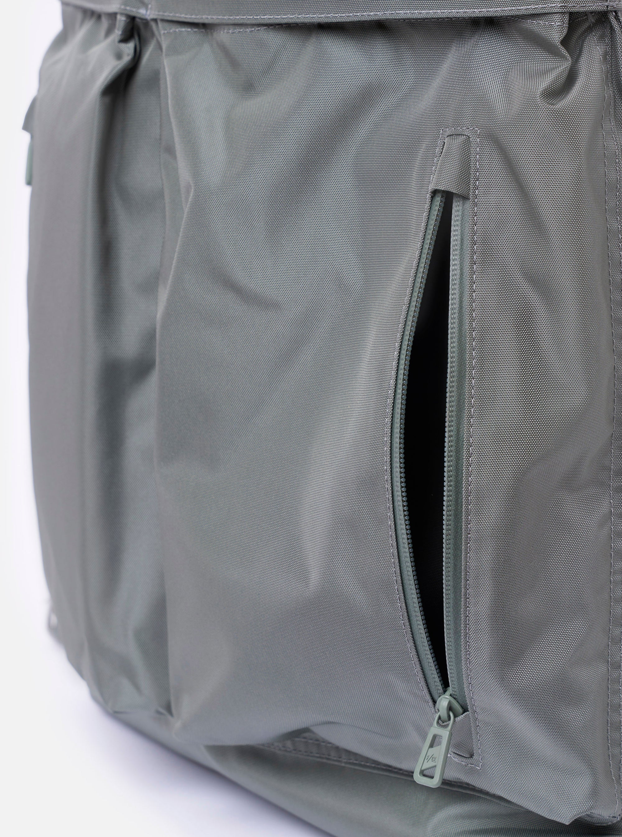 The F/CE. Foliage Green Recycled Nylon 420 re/cor 3-Way Helmet Bag is shown as a flat lay against a plain white background. This foliage green bag features two large front pockets with snap button and Velcro closures, a central vertical zipper for easy access, and a top handle. The design includes adjustable straps that allow it to be worn as a shoulder bag or a backpack.