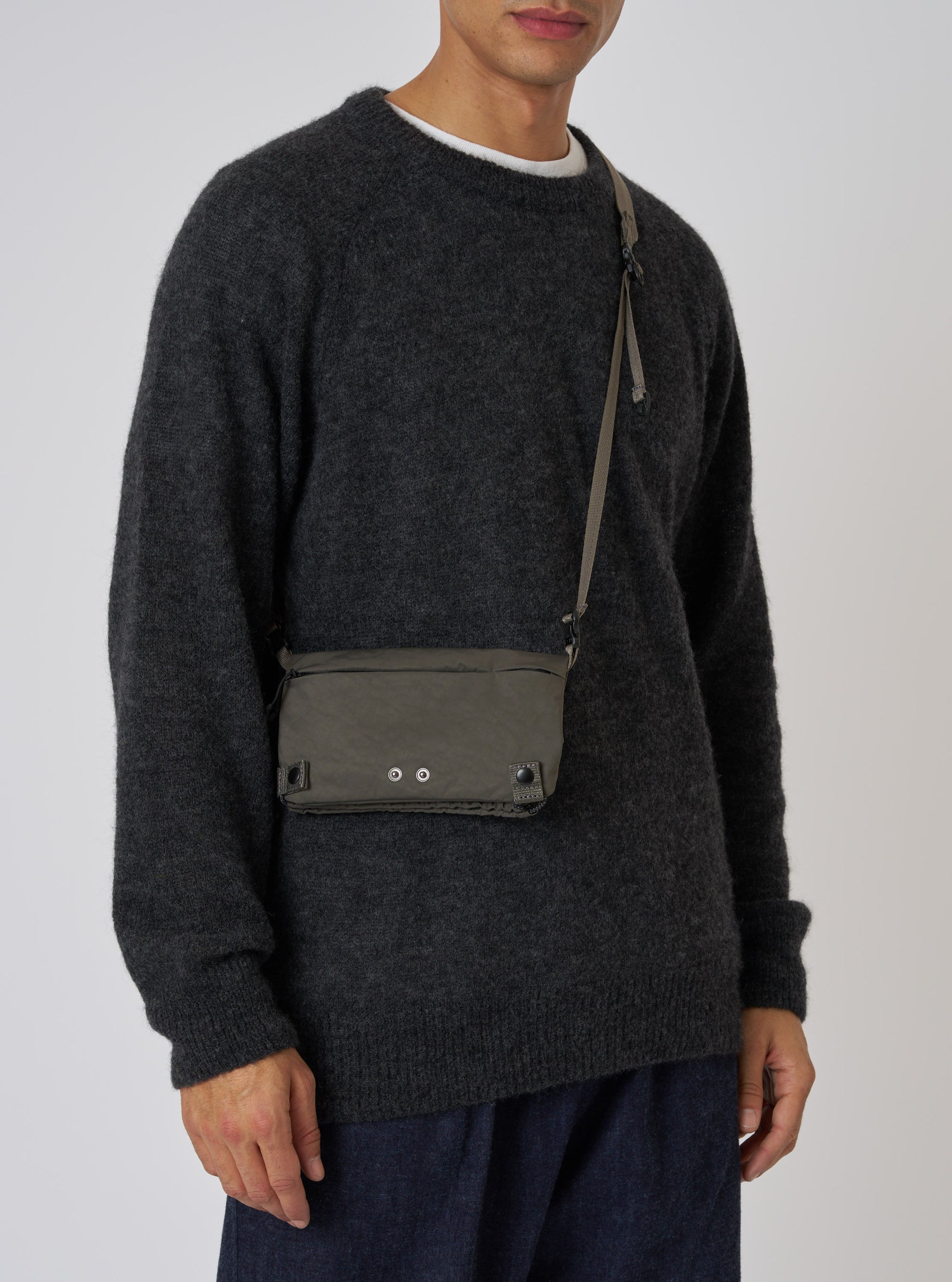 A model showcases the F/CE.® Charcoal Nylon Textured Twill Drawstring Sacoche against a plain backdrop. The bag, with a textured nylon finish, is worn crossbody.
