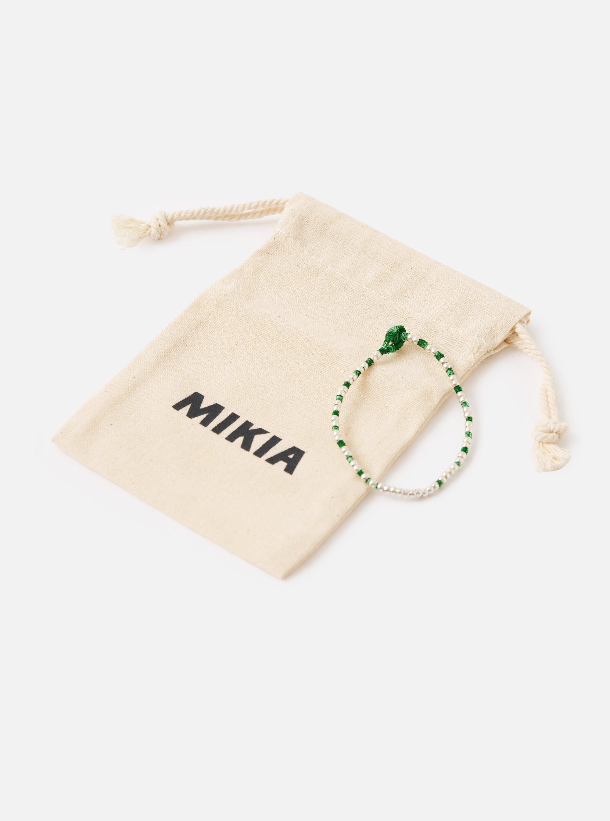The Mikia Malachite Silver Beads Bracelet is displayed next to its branded beige pouch on a white background. The bracelet features green and silver beads.