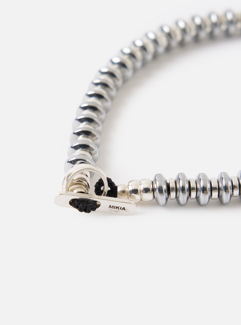 Close-up of the Mikia Hematite Roundel Bracelet on a white surface. The bracelet features metallic hematite beads and a silver clasp with black cord.