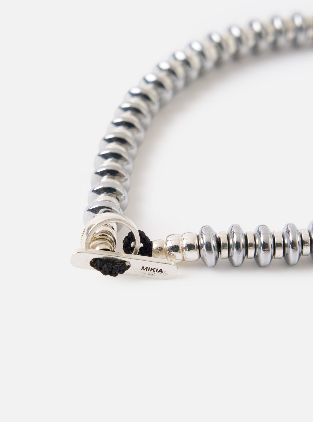 Close-up of the Mikia Hematite Roundel Bracelet on a white surface. The bracelet features metallic hematite beads and a silver clasp with black cord.