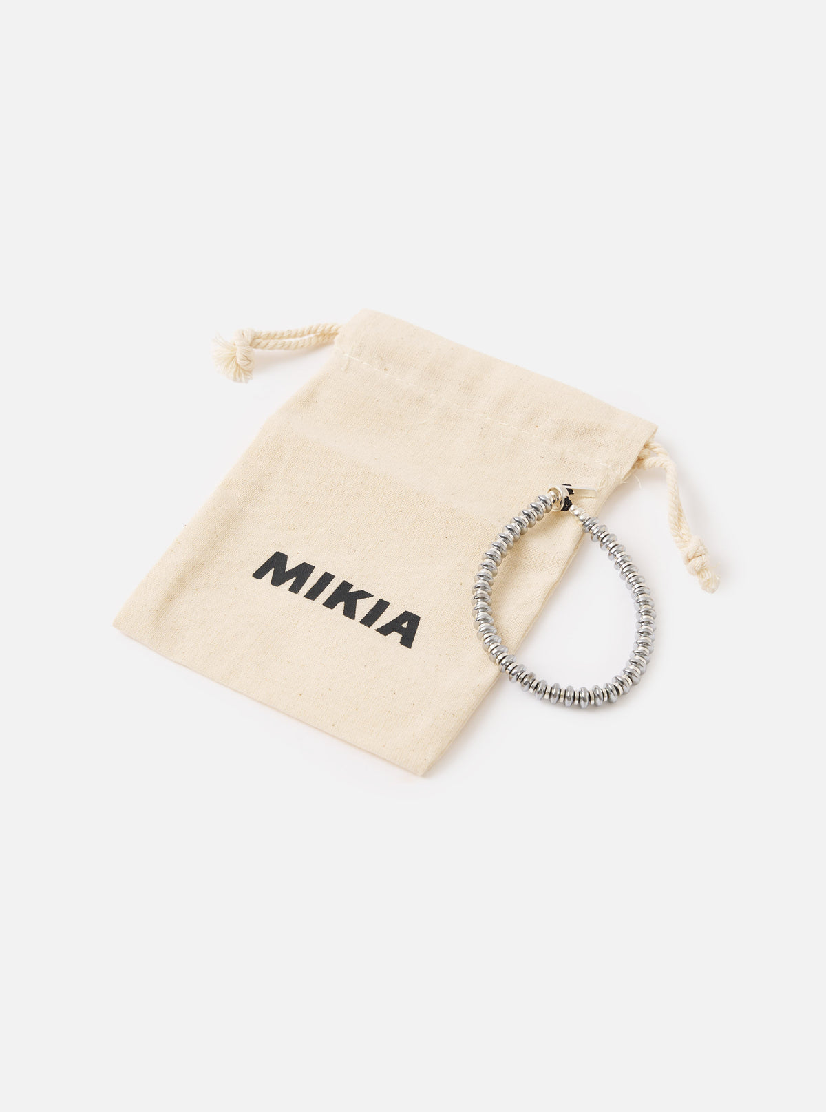 The Mikia Hematite Roundel Bracelet made of gray hematite beads, next to a beige drawstring bag with the Mikia logo, on a white surface.