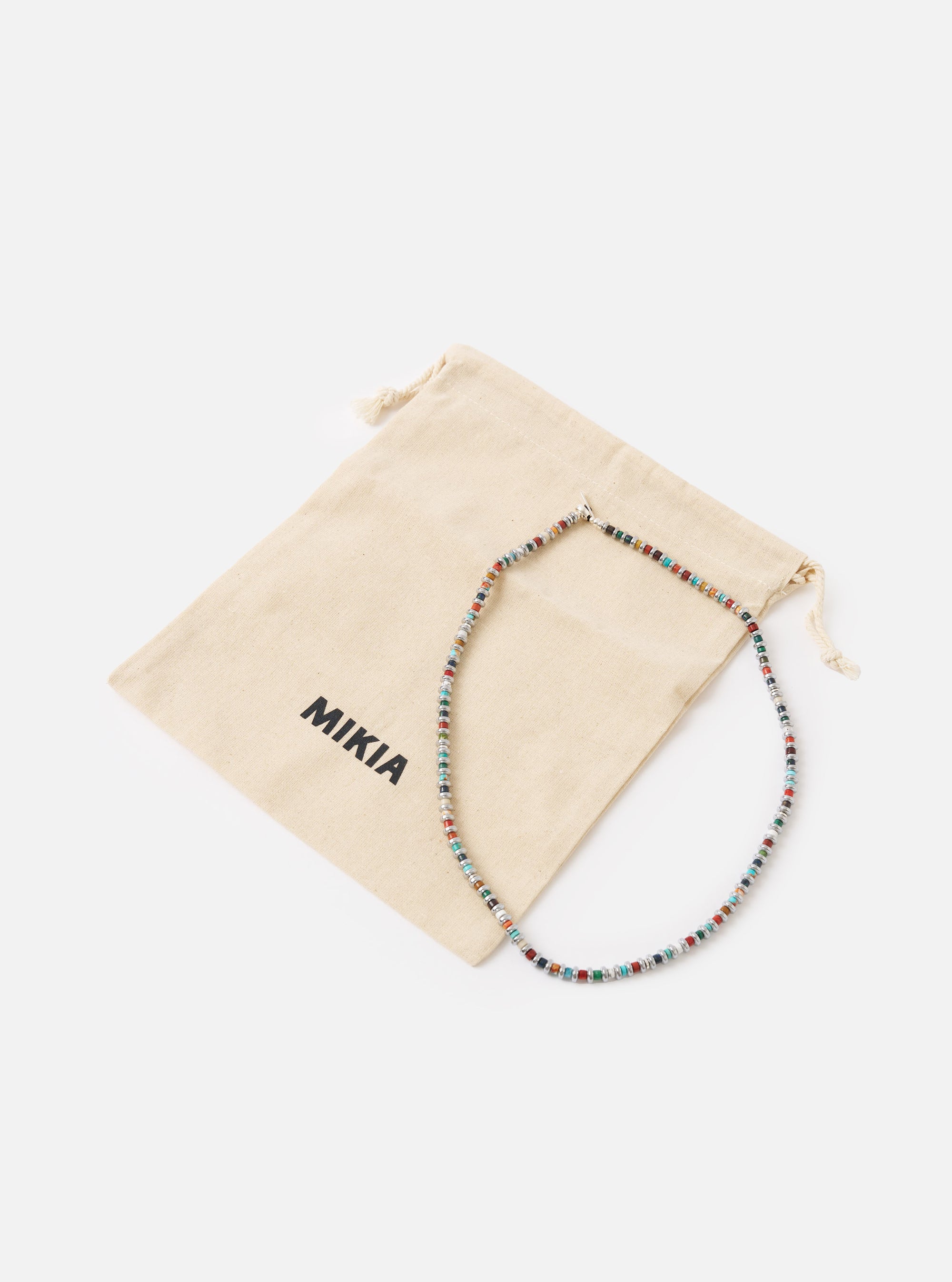 The Mikia Multi Hematite Roundel Necklace on a white surface with its branded beige pouch. The necklace features colorful roundel beads.