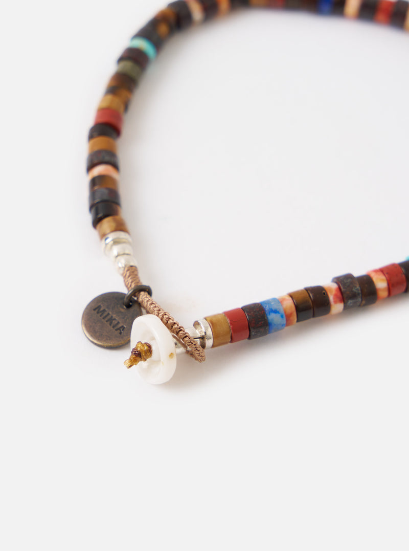 Close-up of a Mikia Brown Multi Heishi Beads Bracelet on a white surface. The bracelet features brown, red, and blue beads with a shell closure and a Mikia logo tag.