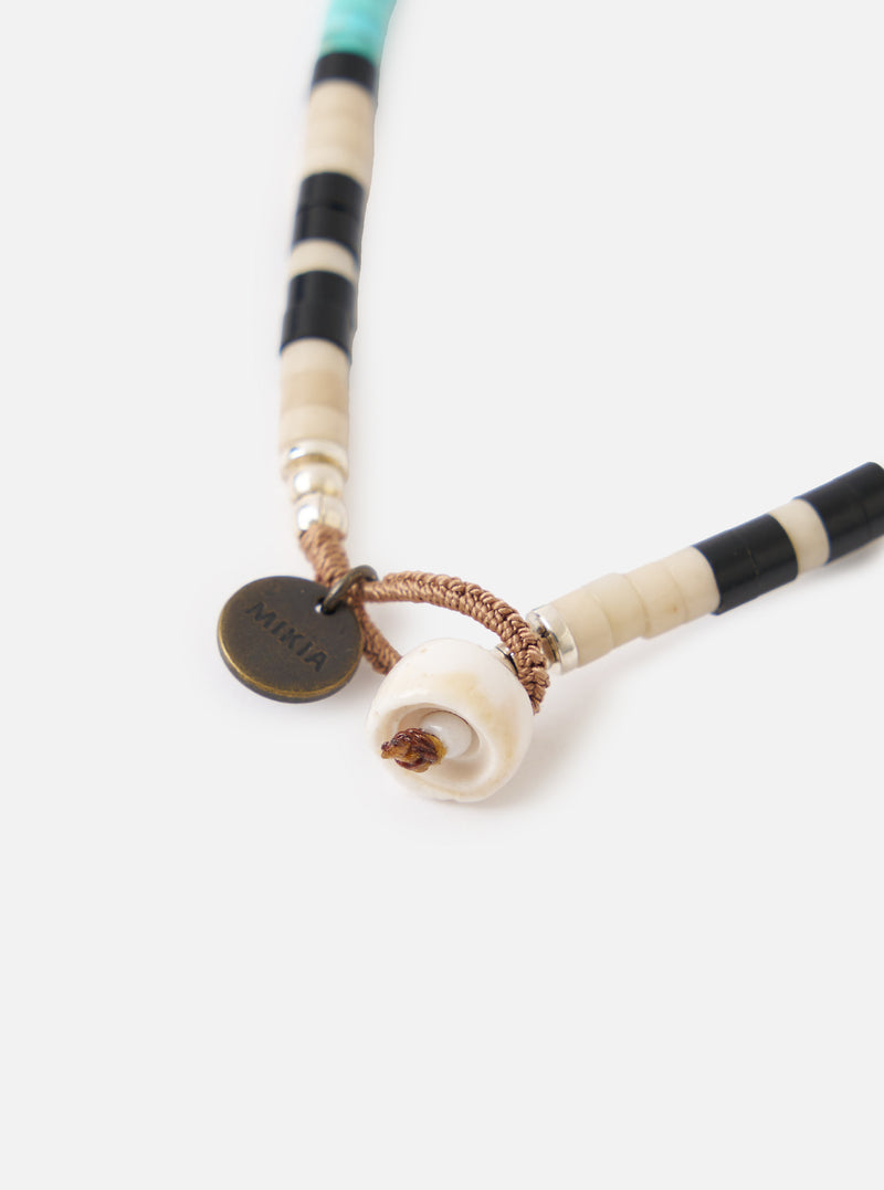 Close-up of the Mikia Fossil Jasper/Turquoise Heishi Beads Bracelet on a white surface. Features turquoise, black, and cream heishi beads, a shell detail, and a Mikia logo charm.