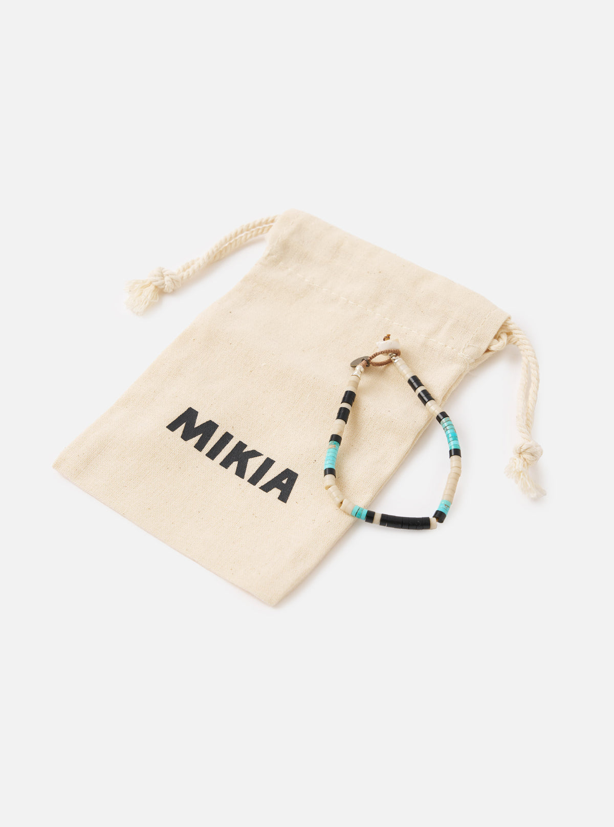 The Mikia Fossil Jasper/Turquoise Heishi Beads Bracelet on a white surface next to its branded dust bag. The bracelet features black, white, and turquoise beads.